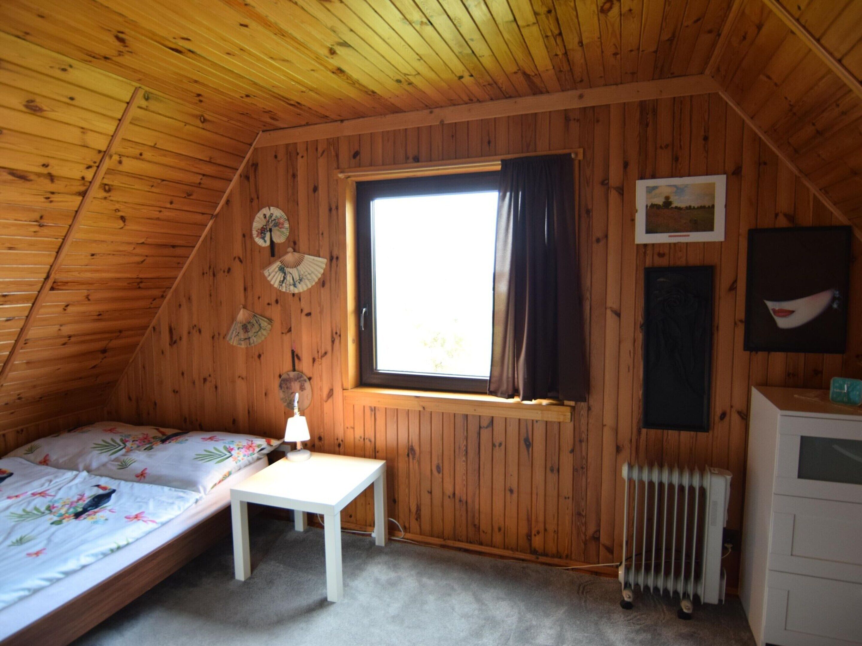 Kodikas ullakkohuone, jonka seinät ovat puuta. Huoneessa on yksi sänky, jossa on kukkakuosiset vuodevaatteet, pieni pöytä, ikkuna, jossa on verhot, ja patteri.