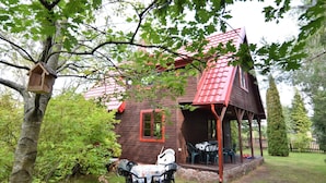 A wooden house with a red roof, a porch with outdoor seating, and a birdhouse on a tree. - Holiday Home With Fireplace and Private Garden (gmina Stężyca)