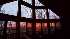 Une pièce dotée de grandes fenêtres, d’un plancher en bois et offrant une vue sur les arbres et le coucher de soleil.