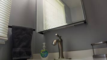Ein Badezimmer mit einem weißen Waschbecken, einem Handtuch, einer Seifenflasche und einem Fenster mit Jalousien.