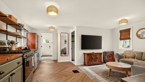 Una cocina moderna con armarios de madera y una estufa, una sala de estar con un sofá y televisor, y un pasillo con un espejo.