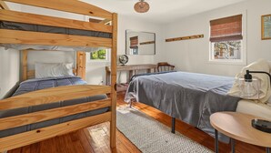 Een kamer met een stapelbed, een bureau, een stoel, een nachtkastje, een lamp en een raam met jaloezieën.