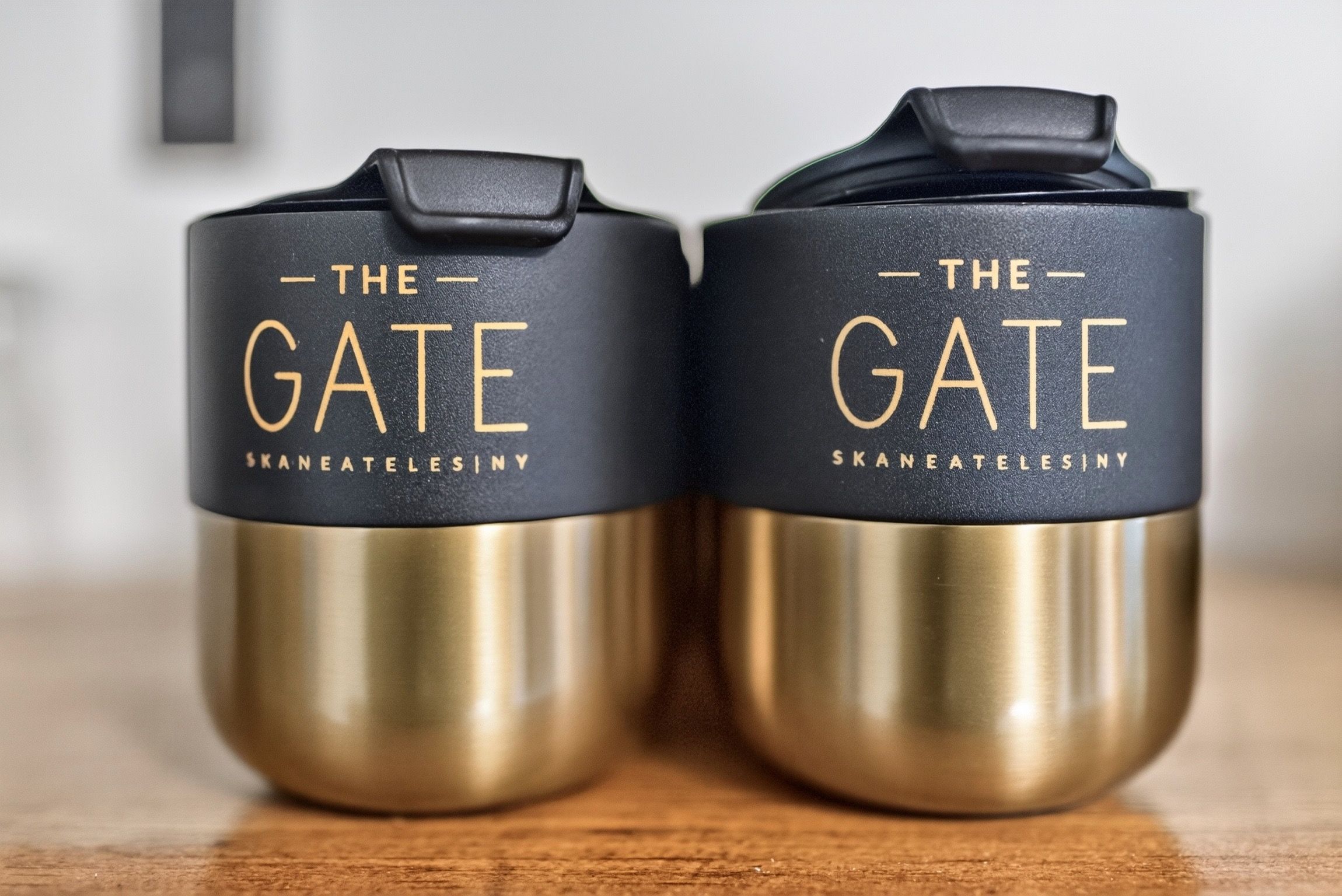 Dos contenedores de acero inoxidable con tapas negras y texto dorado, identificados como 'The Gate Skaneatelesiny'.