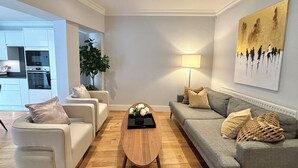 Una sala de estar moderna con un sofá gris, sillones blancos, una mesa de centro de madera y una lámpara de pie. Se ve una cocina al fondo.