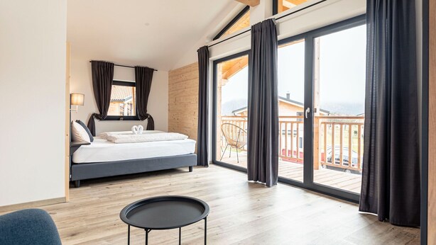 A modern bedroom with a bed, a small round table, and a view of a wooden deck and buildings through large sliding glass doors. - Inzell Chalets With Private Pool (Inzell)