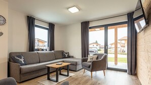 A modern living room with a grey sofa, a small wooden table, and a grey armchair. There is a large window with curtains, a flat-screen TV mounted on the wall, and a clock on the wall. - Inzell Chalets With Private Pool (Inzell)