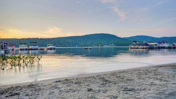 A lakeside view with boats docked at a pier, calm water, and a forested hill in the background. - Cabin w/ Beach Access in Brant Lake: Dogs Welcome! (Brant Lake)