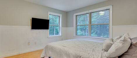 A bedroom with a large bed, two windows, a mounted TV, and wooden flooring.