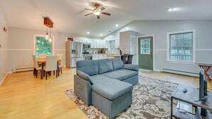 A modern living room with a sectional sofa, dining table, and chairs. A kitchen is visible in the background with white cabinets and appliances. - Cabin w/ Beach Access in Brant Lake: Dogs Welcome! (Brant Lake)