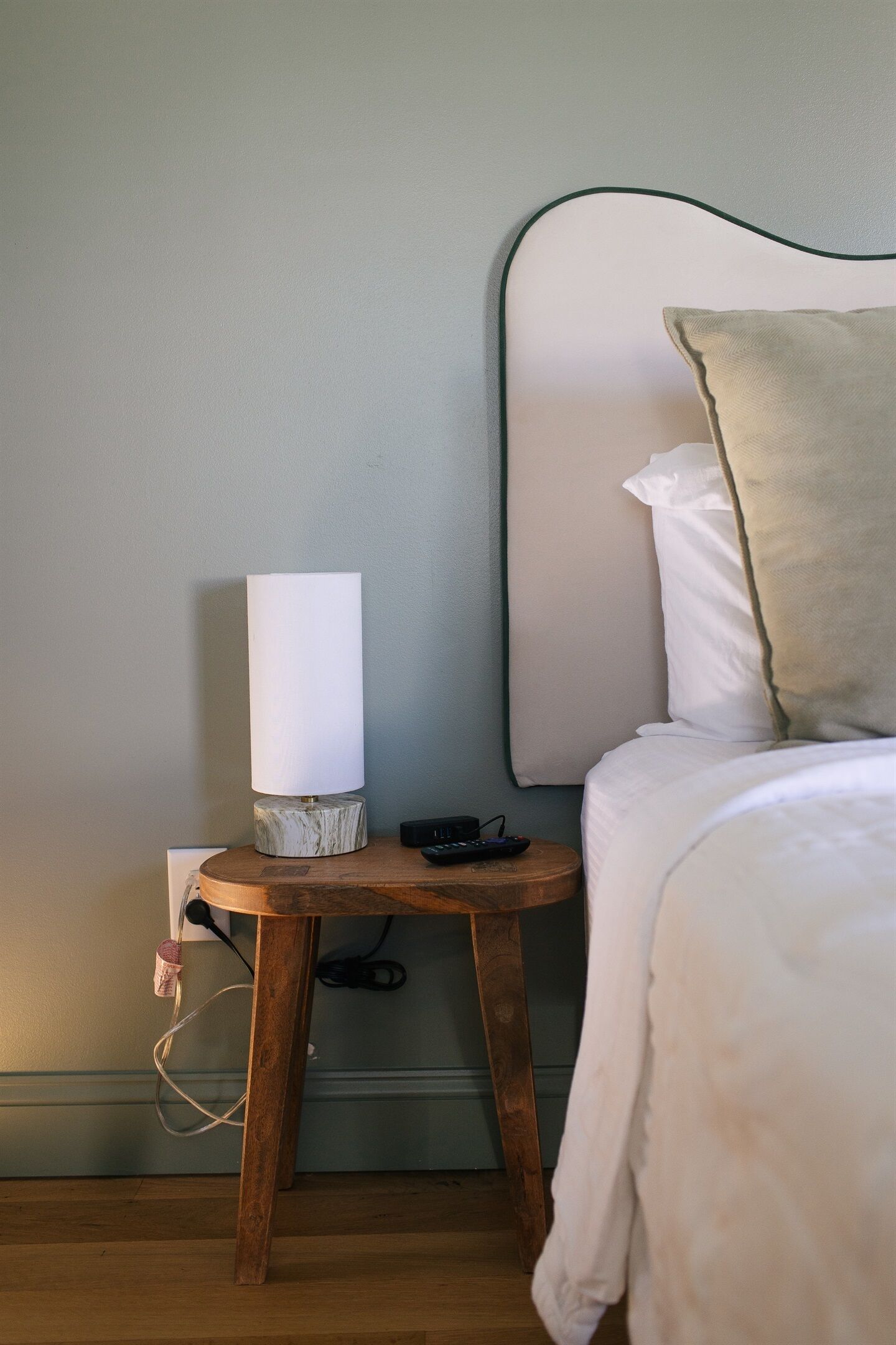 En snyggt bäddad säng med ett vitt täcke och en beige kudde, ett sängbord med en lampa och en fjärrkontroll på golvet bredvid sängen.