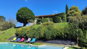 A swimming pool with colorful lounge chairs, a house with a tiled roof, and lush greenery.