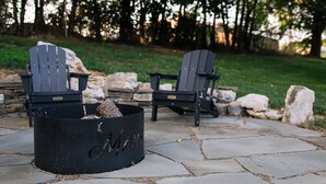 Deux fauteuils Adirondack, séparés par un foyer en métal noir, sont placés sur une terrasse en pierre, avec une pelouse et des arbres en arrière-plan.