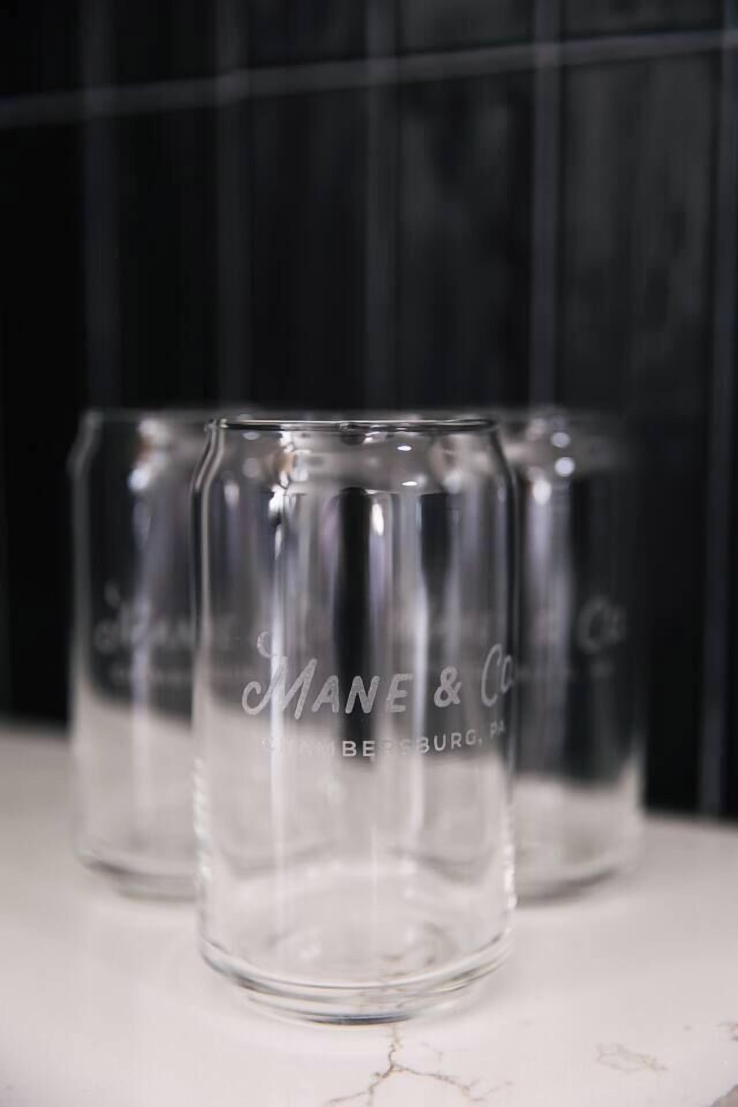 Trois verres à boire en verre transparent, portant la marque « Mane & Co », posés sur une surface en marbre.