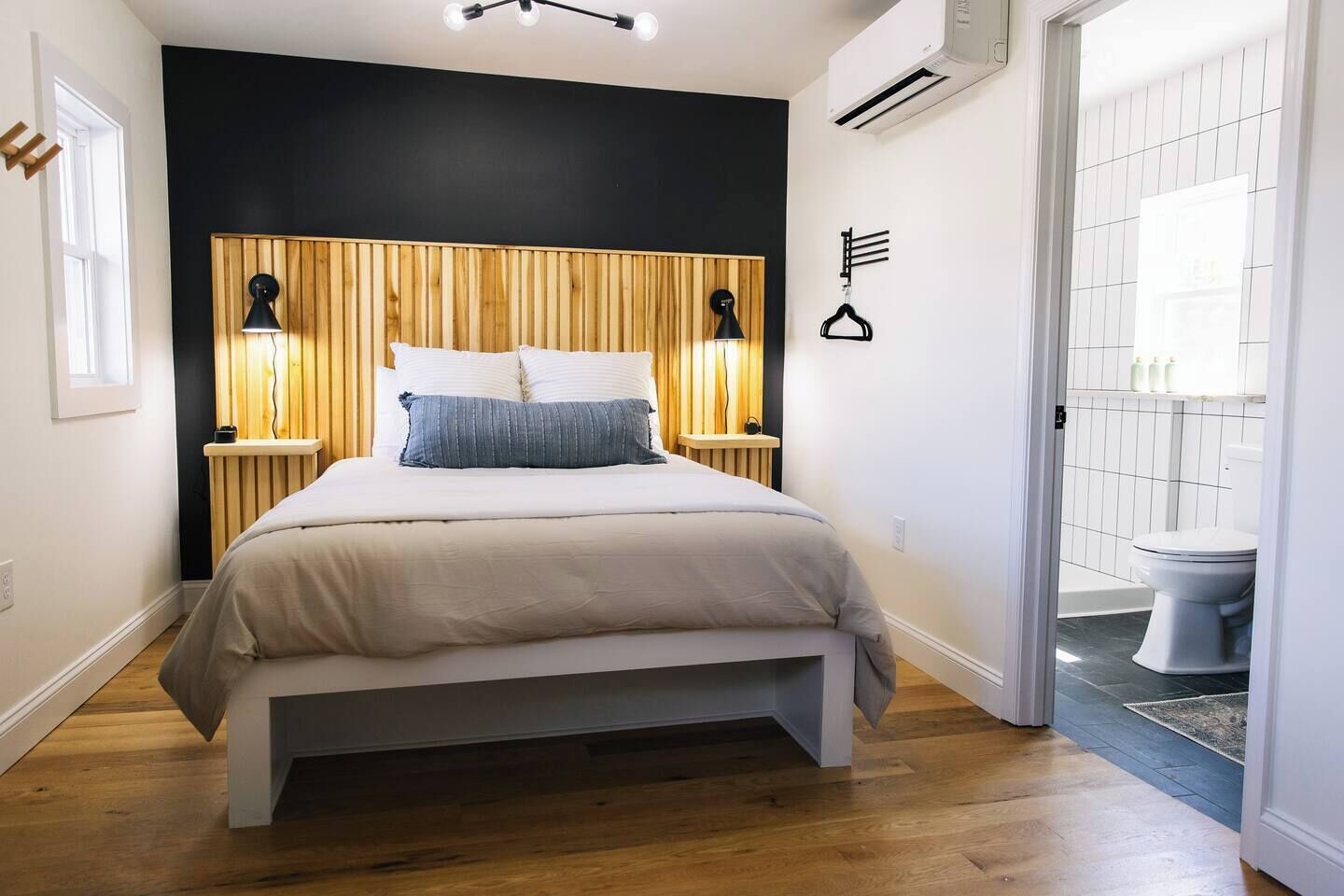 Un dormitorio moderno con cama, lámparas de noche, cabecero de madera y vista al baño.