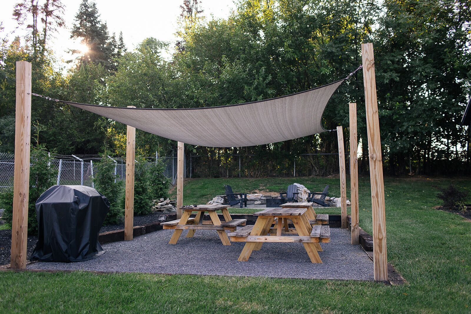Un área de estar al aire libre con techo, mesas de picnic y una parrilla, rodeada por una cerca y árboles.