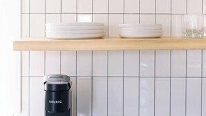 Une cuisine avec des murs recouverts de carreaux blancs, un tableau représentant un paysage encadré, un vase blanc, un récipient blanc, une machine à café noire et une pile d’assiettes blanches.