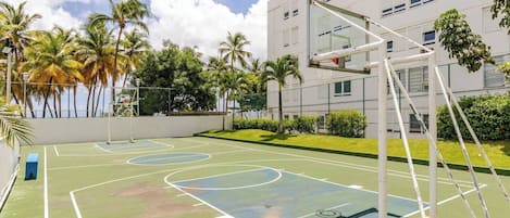 Een buitenlandse basketbalbaan met een net, omringd door palmbomen en een gebouw op de achtergrond.