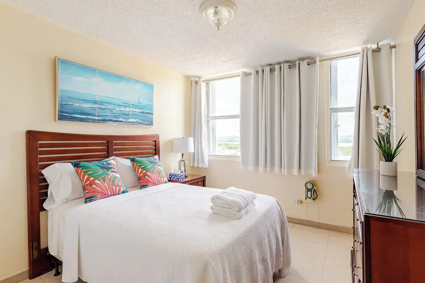 A hotel room with a bed, a nightstand, a lamp, a window with curtains, a painting on the wall, and a plant on the dresser.