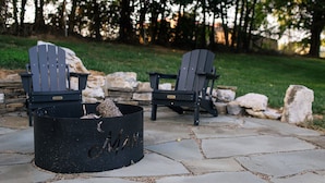 Two Adirondack chairs with a black metal fire pit in between, set on a stone patio with a grassy area and trees in the background.
