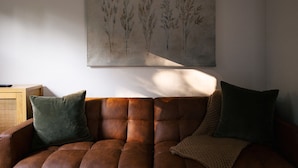 A brown leather sofa with two cushions, a wooden side table, and a wall-mounted painting.