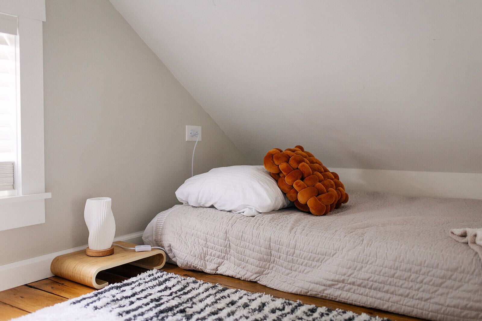 A bedroom with a sloped ceiling, a bed with a white and orange pillow, a white vase on a wooden side table, and a patterned rug.
