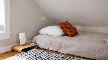 A bedroom with a sloped ceiling, a bed with a white and orange pillow, a white vase on a wooden side table, and a patterned rug.