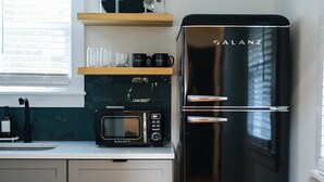 A modern kitchen with a black Galanz refrigerator, a microwave, and a coffee machine on the counter. There are wooden shelves above the counter holding cups and glasses. - Tiny Home with Loft, WI-FI + Free Parking! (Chambersburg)