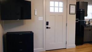 A room with a flat-screen TV mounted on the wall, a white door with a window, a black refrigerator, and a wooden floor.