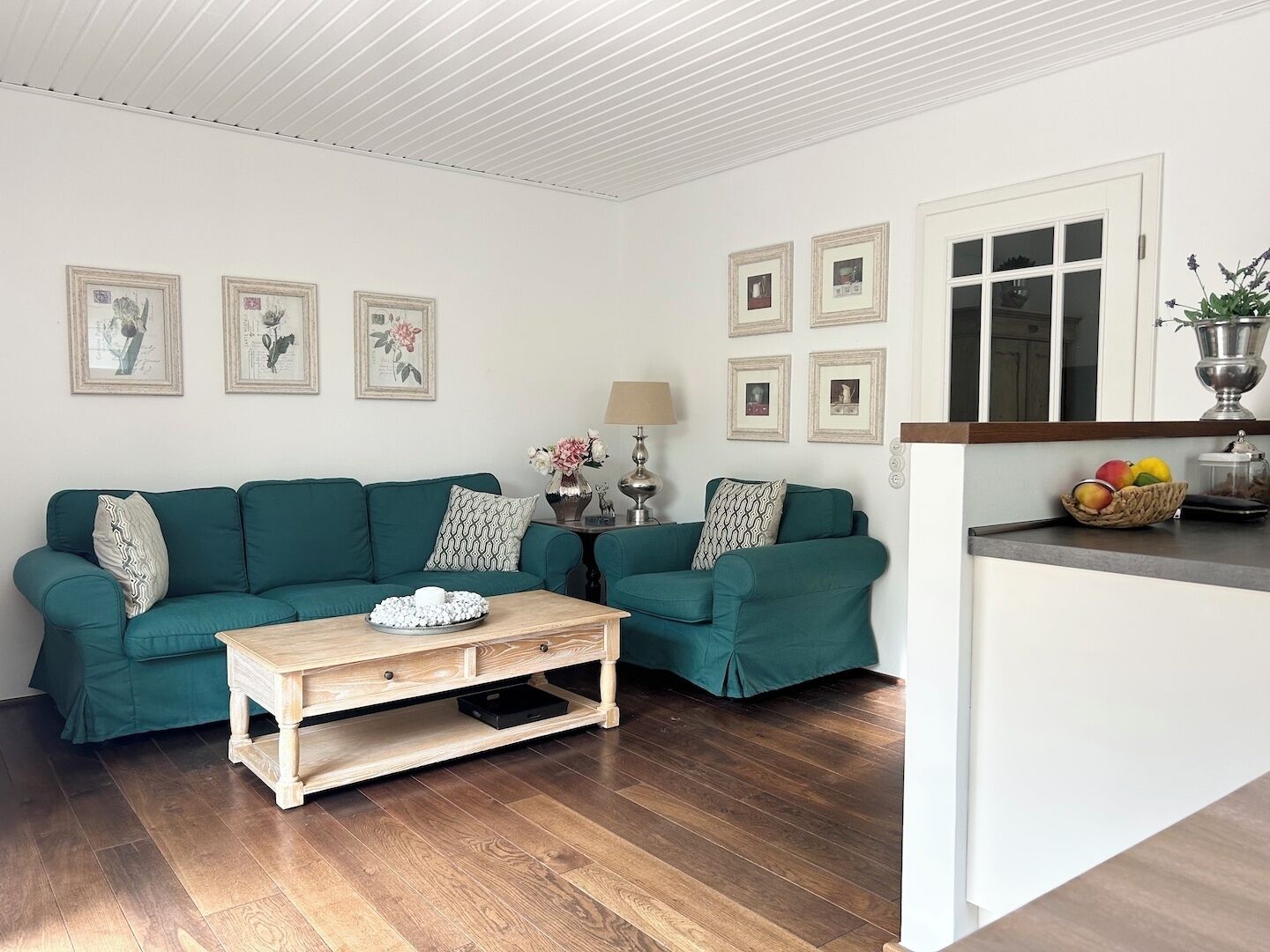 A living room with a teal sofa set, a wooden coffee table, and a kitchen area with a fruit basket.