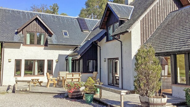 A two-story house with a sloped roof, large windows, and a wooden deck with outdoor seating. - A magnificent property in a secluded position in Evanton. (Evanton, near Dingwall)