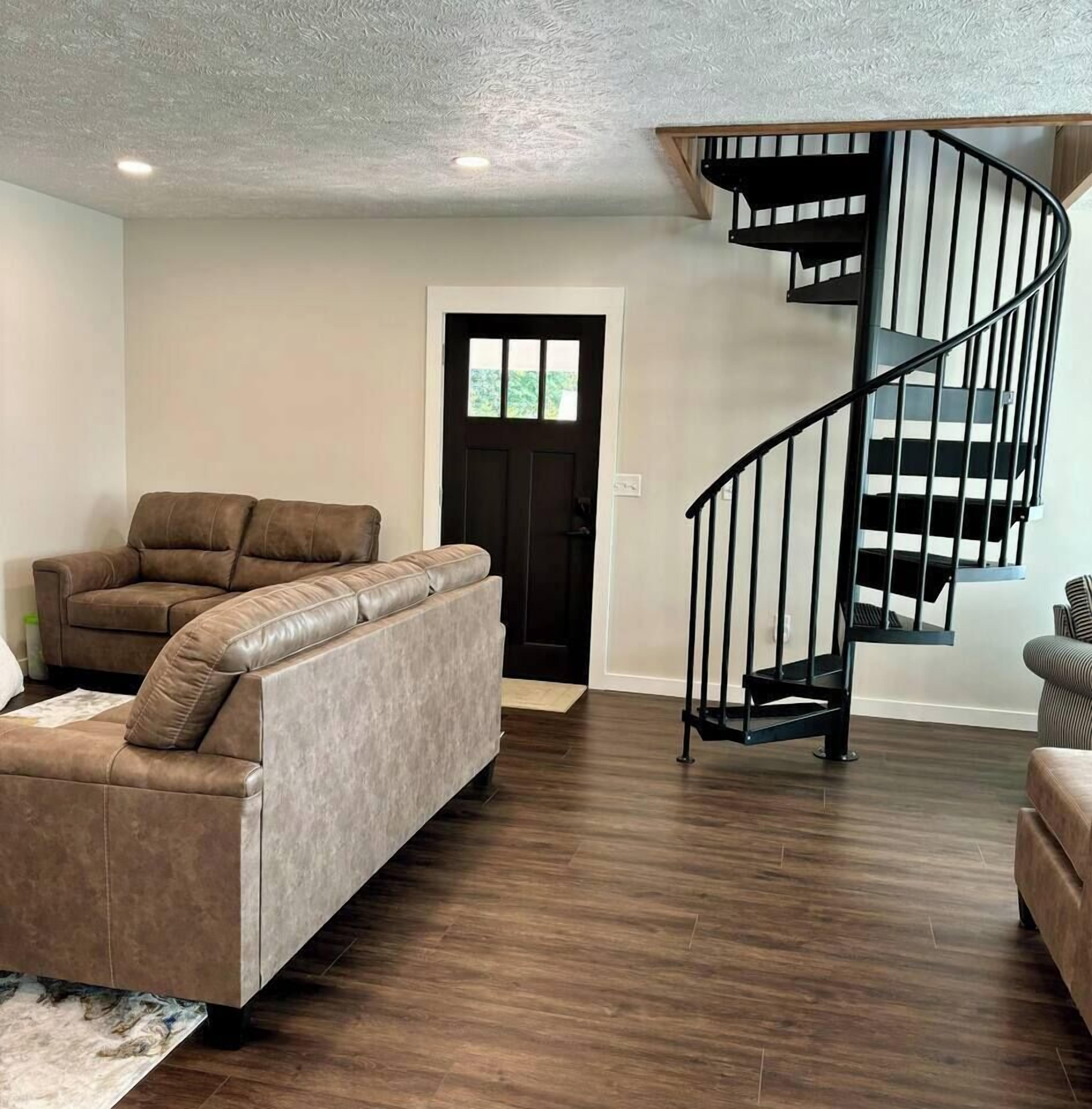 A living room with a brown leather sofa, a wooden staircase, and a dark door.