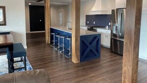 A modern kitchen with dark wood flooring, a central island with stools, stainless steel appliances, and a blue accent wall. - Pristine Home Perfect for Family Bay Views Luxe Chef’s Kitchen Ludington Park (Escanaba)