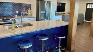 A modern kitchen with a granite countertop, stainless steel appliances, and blue bar stools. - Pristine Home Perfect for Family Bay Views Luxe Chef’s Kitchen Ludington Park (Escanaba)