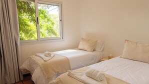 Una habitación con dos camas, cada una con cubrecama y almohadas blancas, y un ventanal con cortinas.