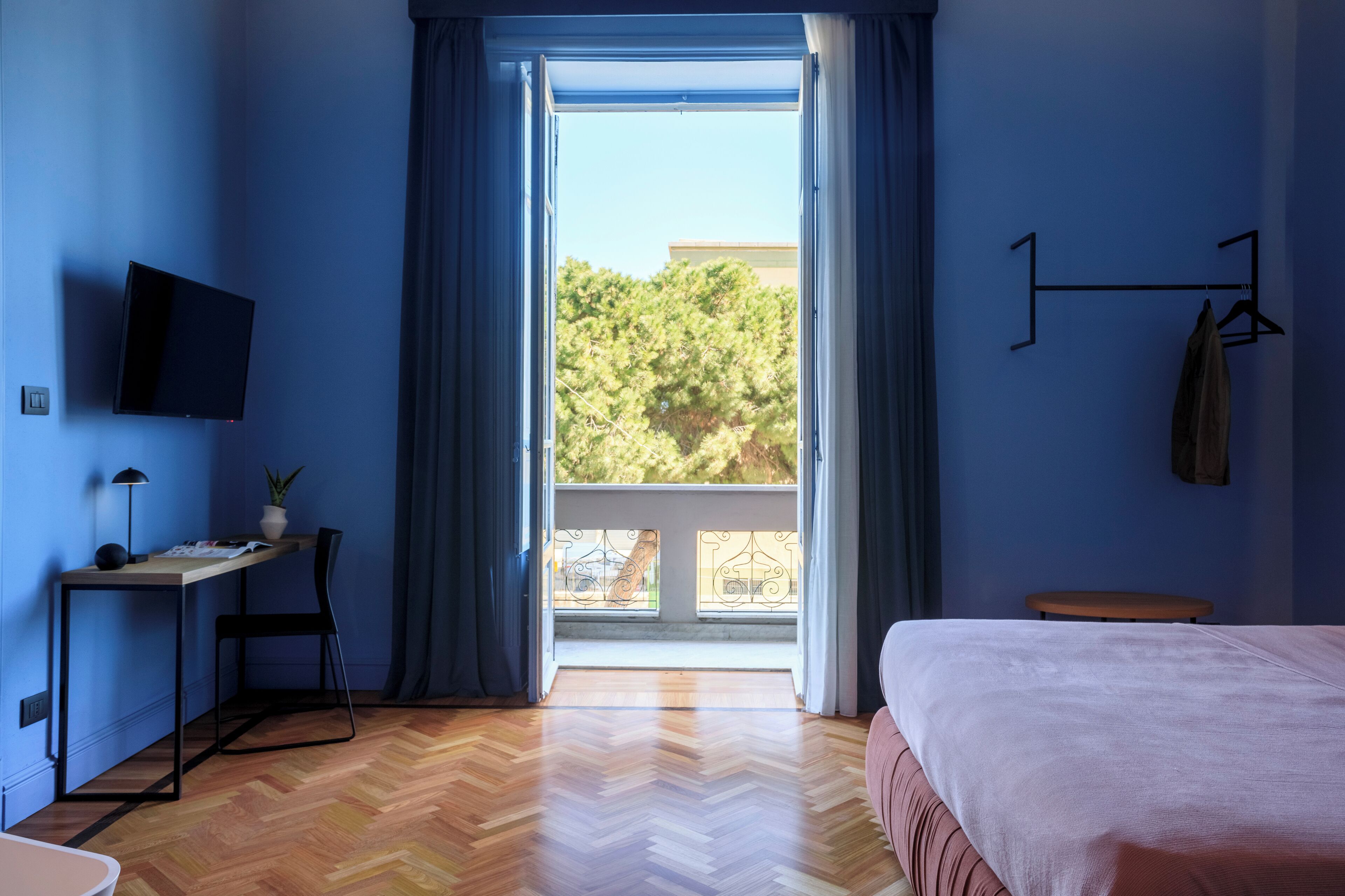 A bedroom with a bed, a desk with a chair, a TV mounted on the wall, and a view of trees through the window.
