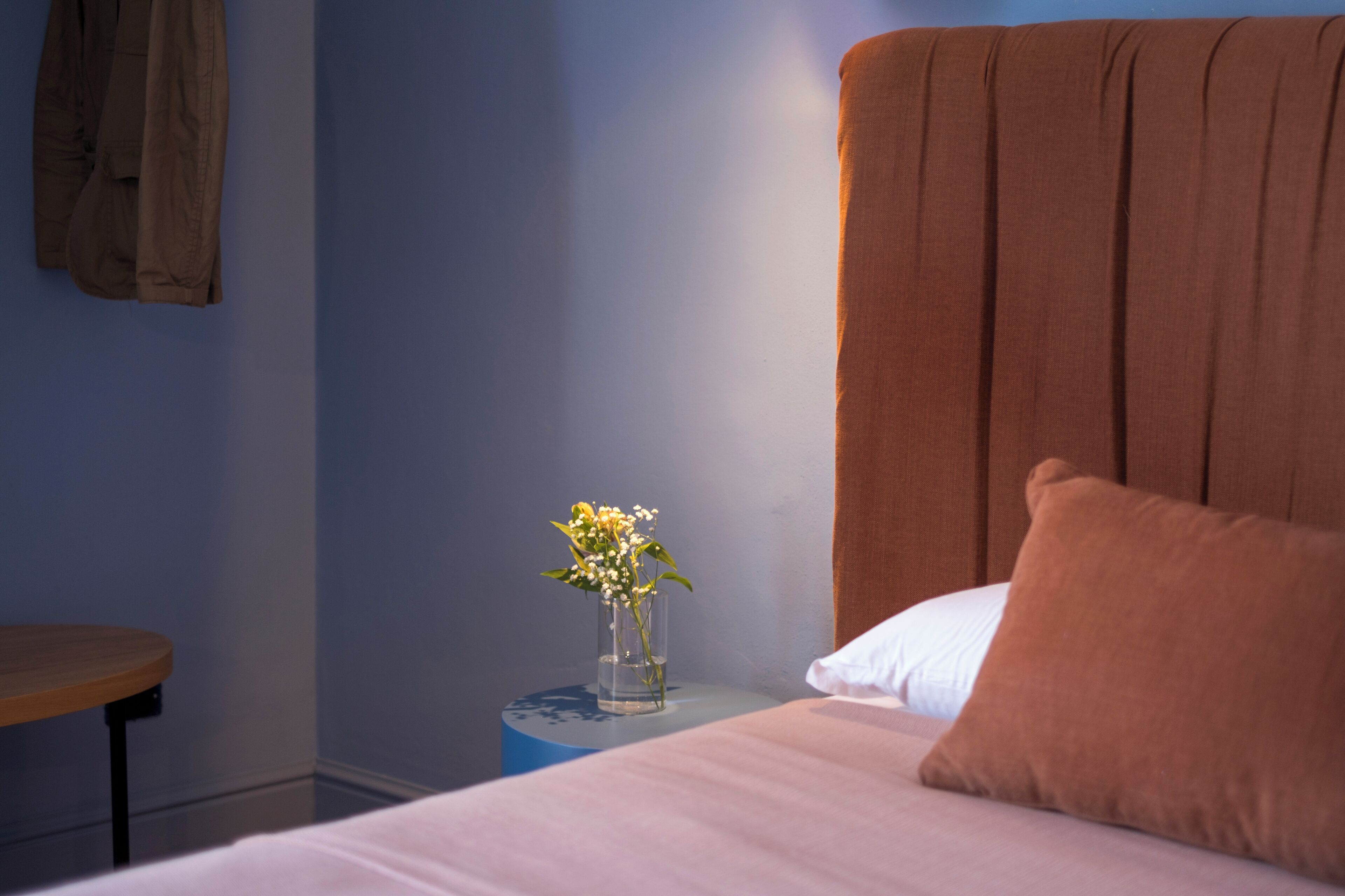 A bedroom with a bed, a nightstand with a vase of flowers, a wooden chair, and a brown headboard.