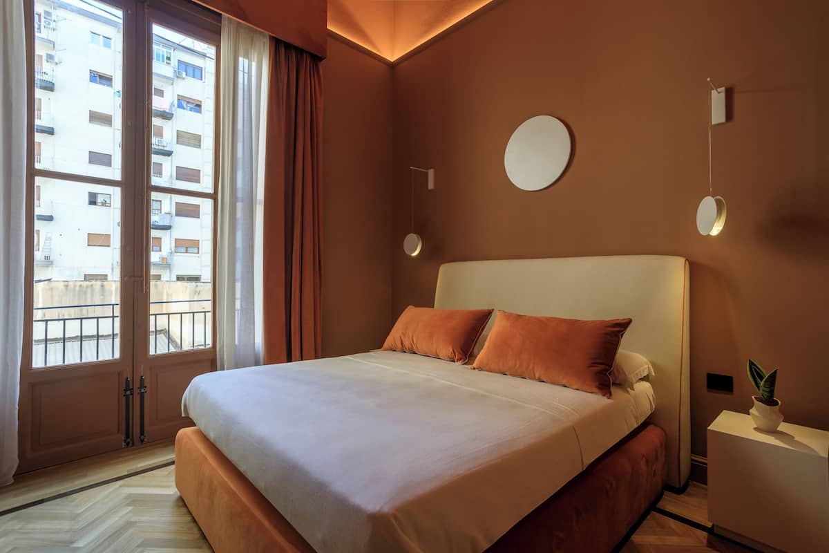 A modern hotel room with a large bed, a view of the city through the window, and a round mirror on the wall.