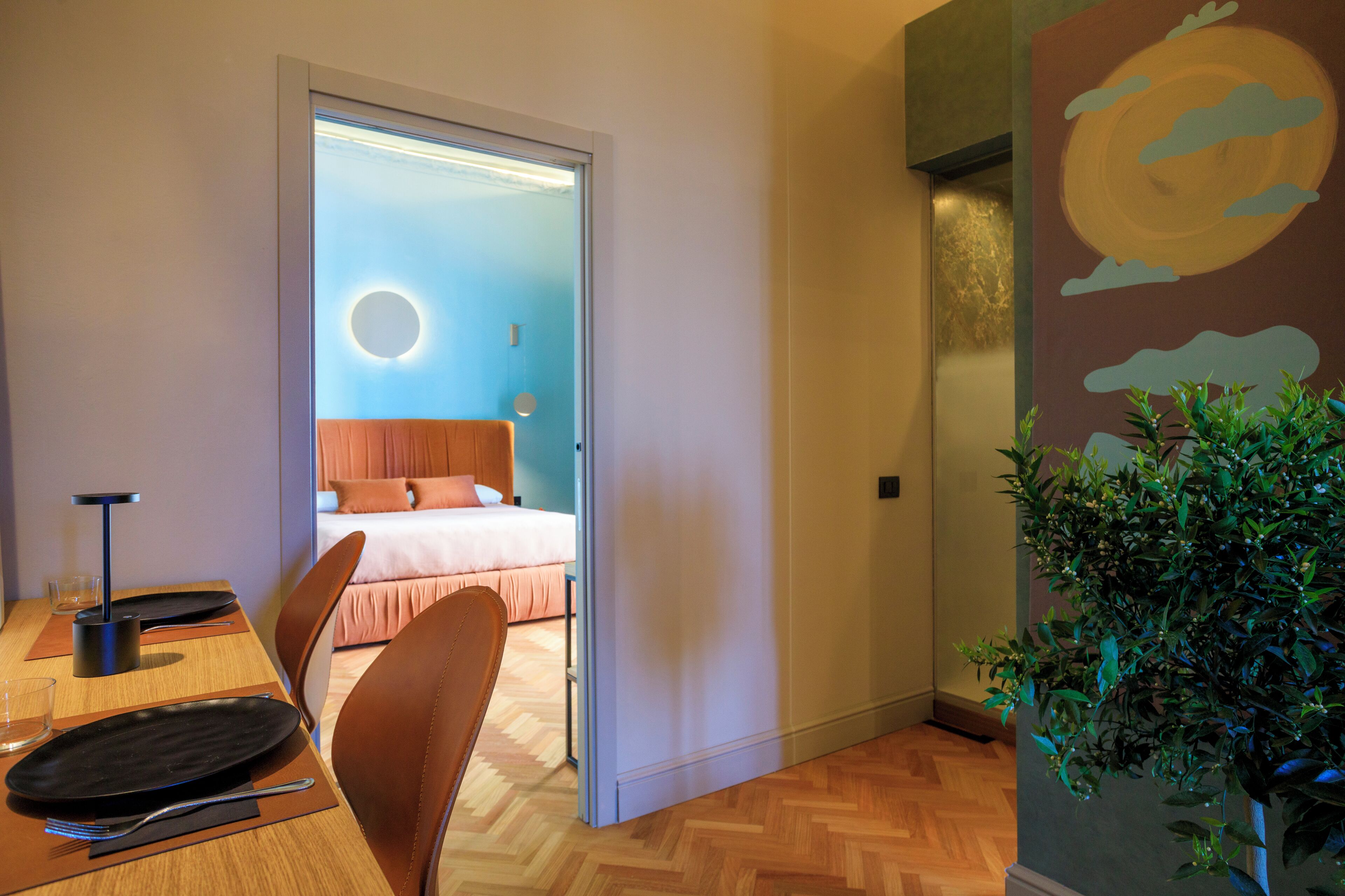 A modern hotel room with a wooden floor, a bed with a circular light fixture, a dining area with a table and chairs, and a wall-mounted plant.