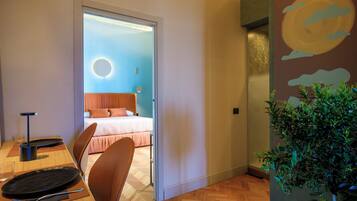 A modern hotel room with a wooden floor, a bed with a circular light fixture, a dining area with a table and chairs, and a wall-mounted plant.