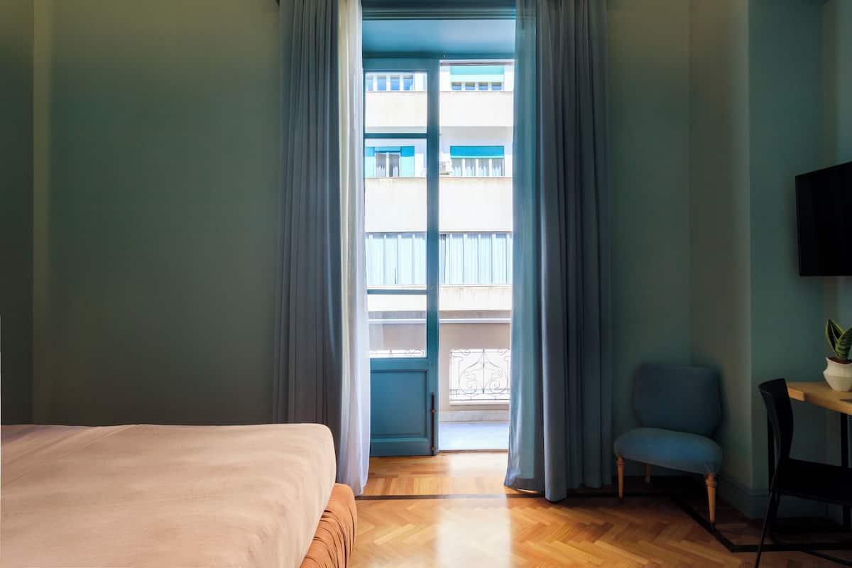 A hotel room with a bed, a chair, a small table with a plant, and a view of a building through a window with curtains.
