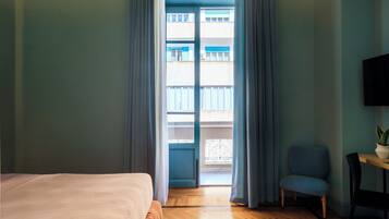 Ein Hotelzimmer mit einem Bett, einem Sessel, einem kleinen Tisch mit Pflanze und Blick auf ein Gebäude durch ein Fenster mit Vorhängen.