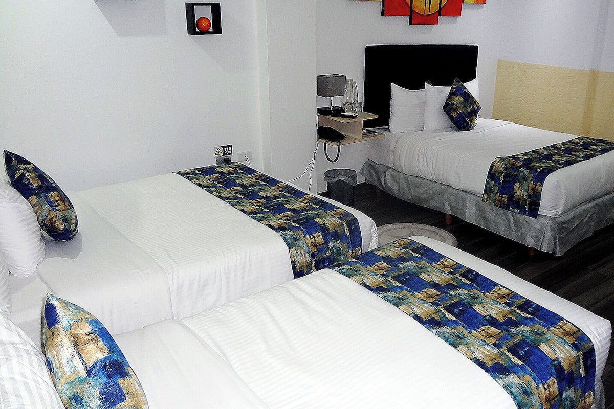 A hotel room with two single beds, each with a blue and gold patterned bedspread, white pillows, and a small bedside table with a lamp and a phone.
