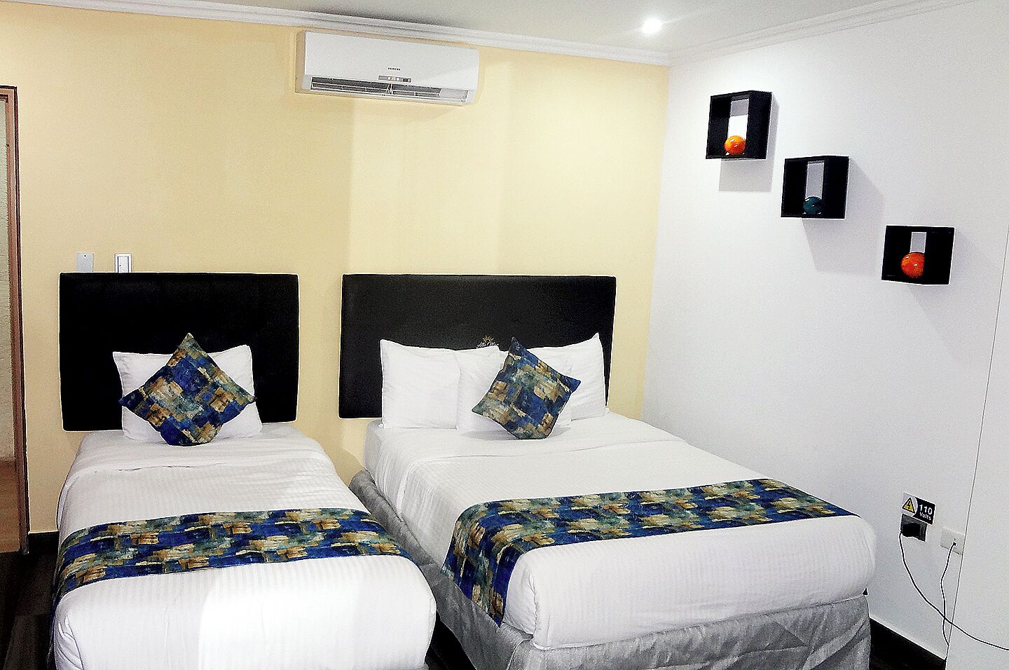 Une chambre d’hôtel avec deux lits, chacun doté d’une tête de lit noire et de draps blancs. Un appareil de climatisation est fixé au mur au-dessus des lits.