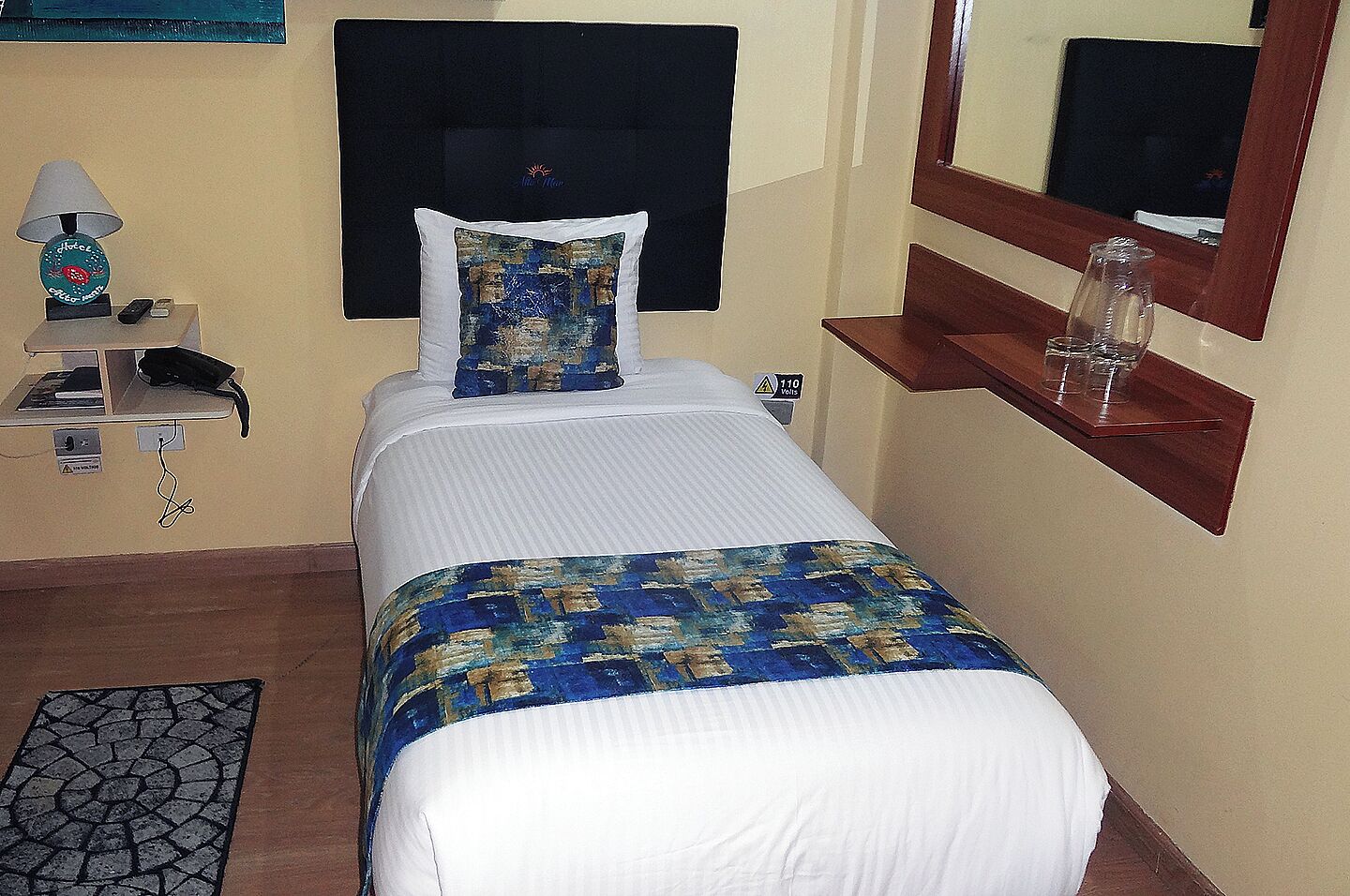 A hotel room with a bed, a nightstand with a lamp, a phone, and a mirror.