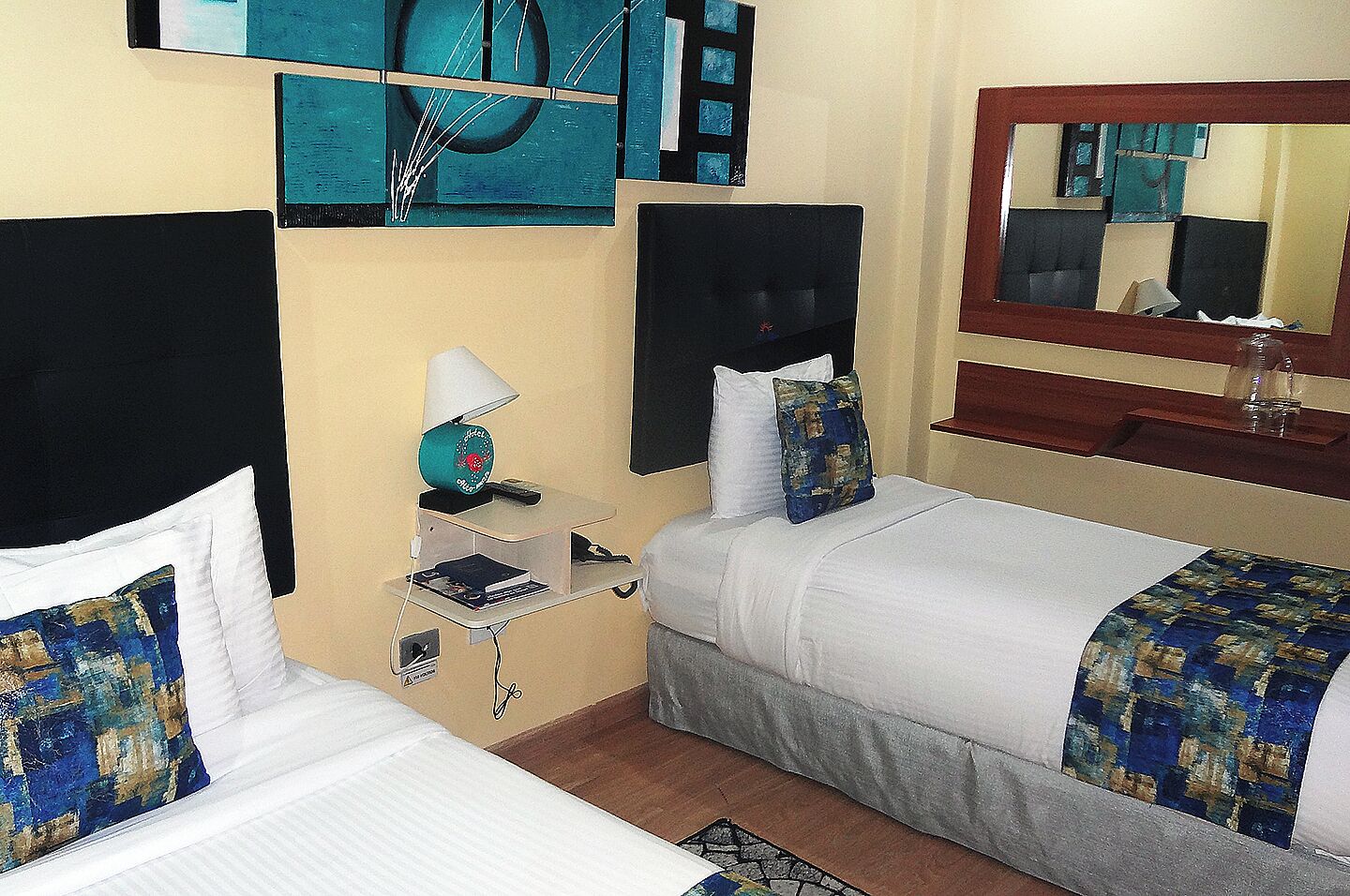 Habitación de hotel con dos camas, un escritorio con lámpara, un espejo y arte abstracto en la pared.