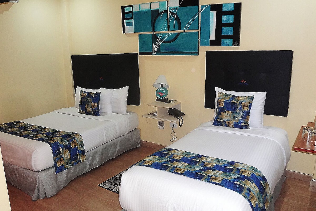 A hotel room with two single beds, each with a white comforter and blue patterned bedspreads. There is a small bedside table with a lamp and a phone. The walls are adorned with abstract art.