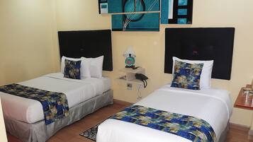 A hotel room with two single beds, each with a white comforter and blue patterned bedspreads. There is a small bedside table with a lamp and a phone. The walls are adorned with abstract art.