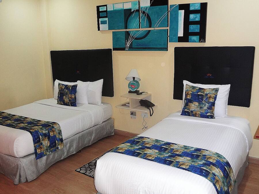A hotel room with two single beds, each with a white comforter and blue patterned bedspreads. There is a small bedside table with a lamp and a phone. The walls are adorned with abstract art.