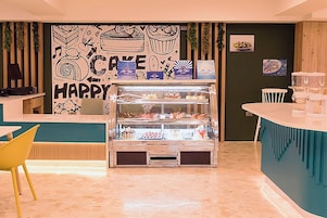 Un mostrador de panadería con una variedad de productos horneados exhibidos en una vitrina, un menú en una pizarra y una pared con un mural con temática de pasteles.