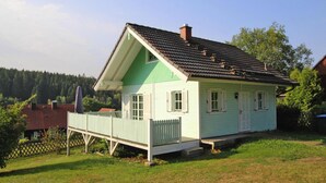Ein kleines, eingeschossiges Haus mit weißer Fassade und grünen Verzierungen, mit einer Veranda und einem eingezäunten Garten.